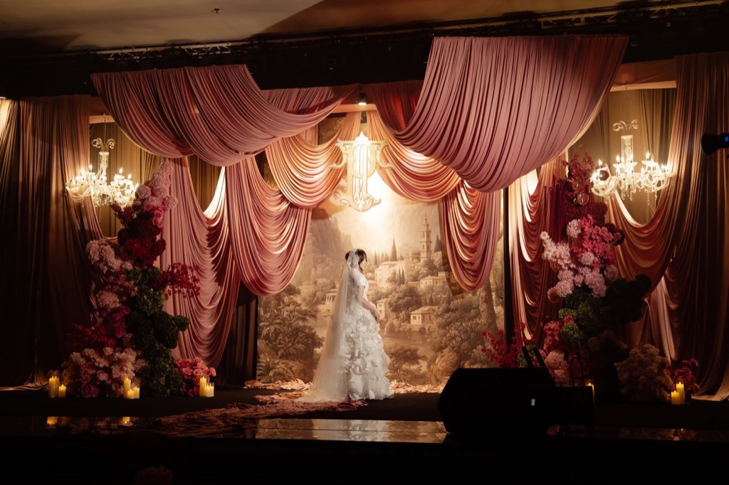 A Rose Affair — Cô dâu đứng giữa sân khấu rèm lụa hồng xếp tầng, hai đèn chùm pha lê, tranh phong cảnh cổ điển và hai trụ hoa hồng bên ánh nến