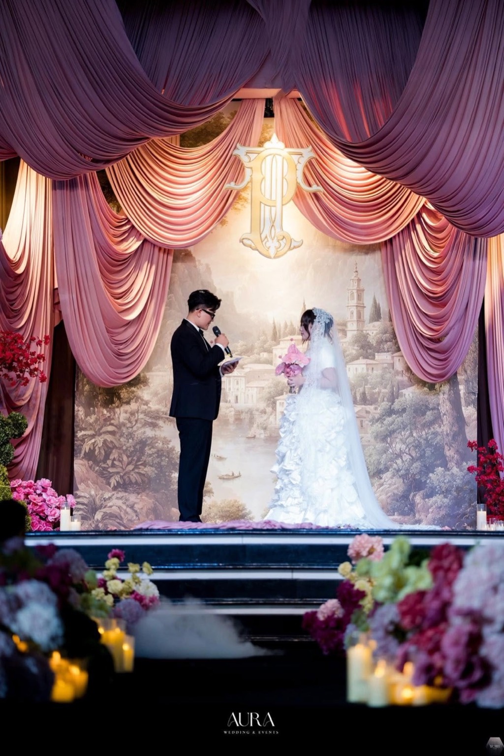 Cô dâu chú rể trao lời thề trên sân khấu rèm lụa hồng, monogram vàng và tranh phong cảnh, tiền cảnh hoa và nến