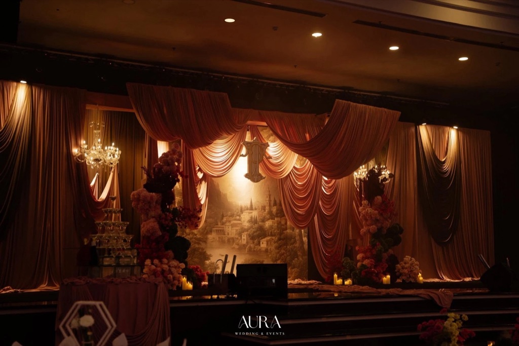 A Rose Affair — Toàn cảnh sân khấu rèm hồng trong sảnh tối, tháp ly champagne, monogram mạ vàng, tranh phong cảnh và trụ hoa hai bên