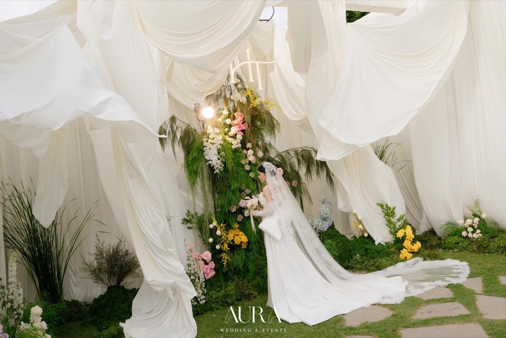 Cô dâu giữa những dải vải trắng bay nhẹ, phía sau là cụm hoa cỏ xanh và monogram HT
