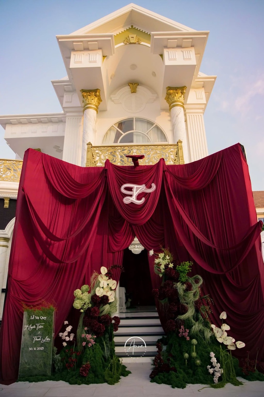 Cổng vu quy trước biệt thự trắng: drapery đỏ burgundy rủ nhiều tầng, monogram cưới và hai khối hoa garden form trên nền rêu