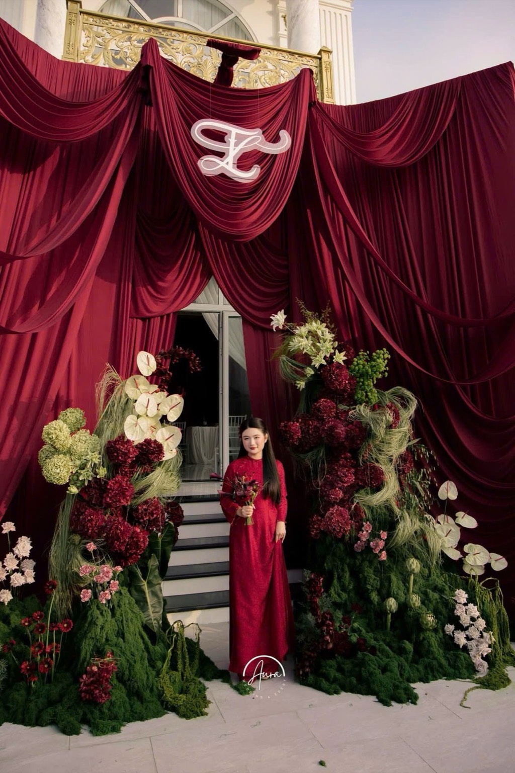 Cô dâu áo dài đỏ giữa cổng drapery burgundy và hai khối hoa garden form