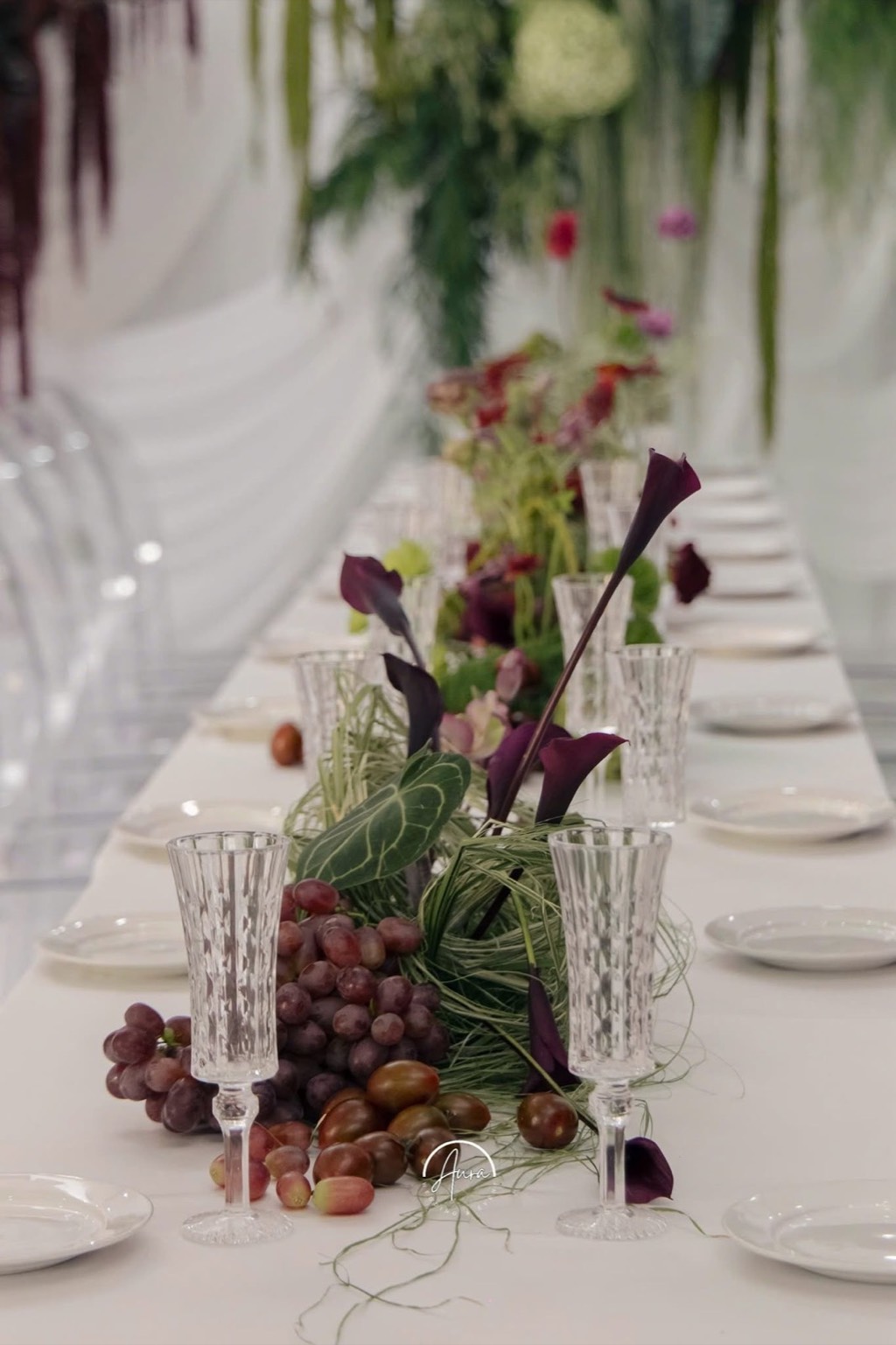 Bàn tiệc feast-style: ly pha lê, nho, mận và hoa rum tím trên khăn trắng