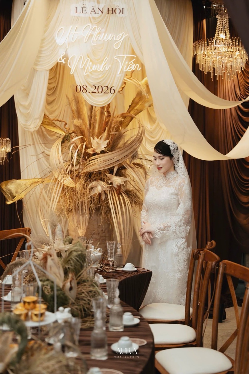 Bàn tiệc khăn nâu với ly pha lê, hoa khô và ghế gỗ trước backdrop lụa kem và đèn chùm pha lê