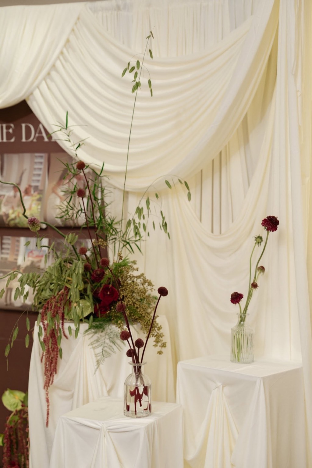 Lụa trắng ngà buông rủ, bục phủ vải với bình hoa đỏ đậm, dền đỏ rủ và nhánh lá xanh
