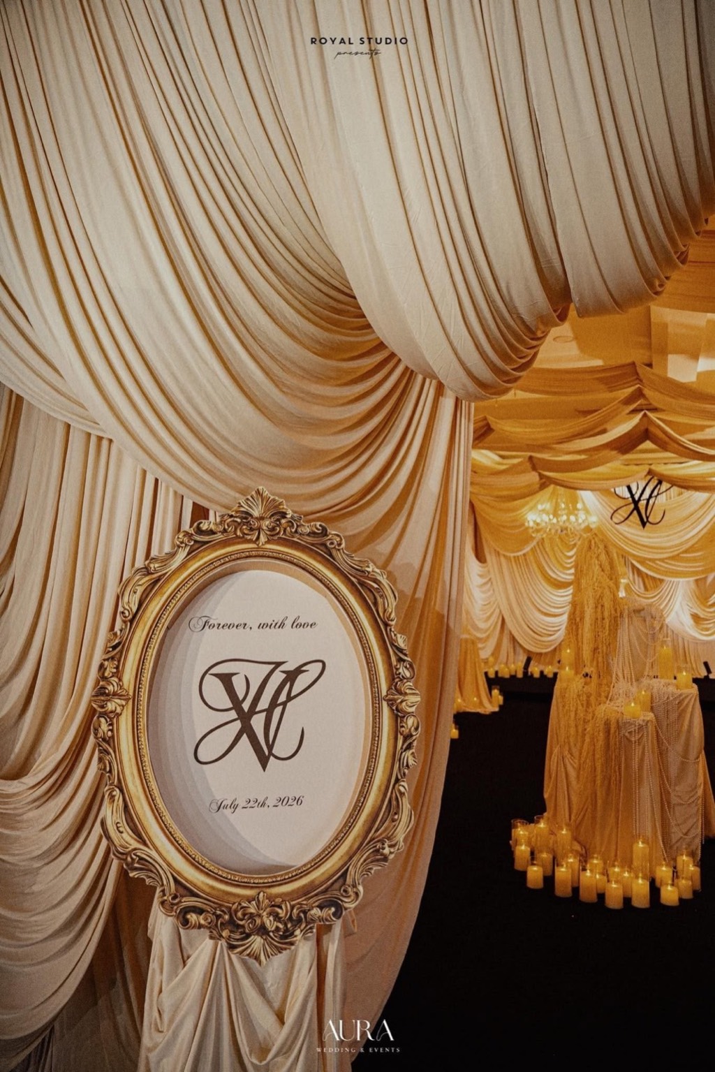 Bảng chào khung oval mạ vàng giữa vòm lụa ngà, phía xa là sân khấu monogram và nến