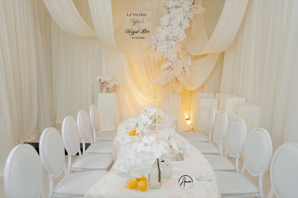 Toàn cảnh bàn gia tiên dài phủ khăn trắng với lan hồ điệp, chanh vàng, hàng ghế lưng oval trắng và backdrop vải kem buông rũ cùng thác lan treo