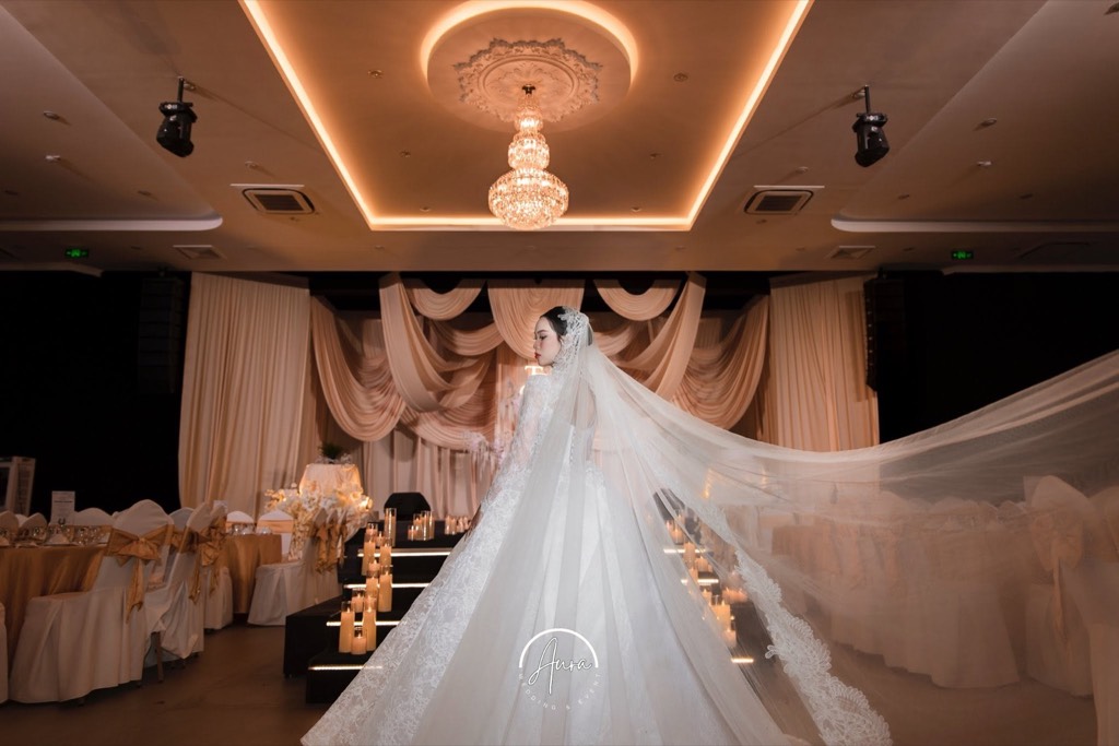 Cô dâu với tà voan bay giữa sảnh tiệc, sân khấu rèm champagne xếp tầng, đèn chùm pha lê và bậc thang viền nến