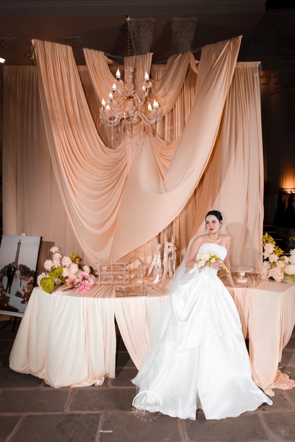 Bàn chào phủ vải champagne dưới đèn chùm nến, rèm vắt cao và hoa tươi hai bên, cô dâu ngồi trước bàn
