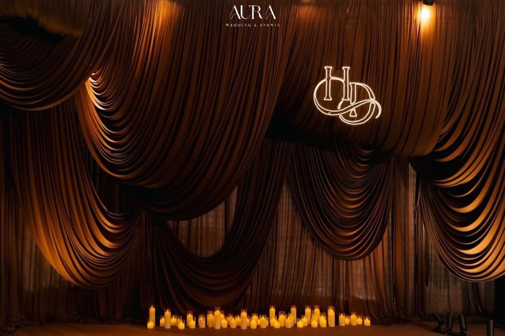 The Amber Hour — Phông rèm nâu xếp rủ nhiều tầng được đánh đèn vàng hổ phách, monogram HD và hàng nến dưới chân sân khấu