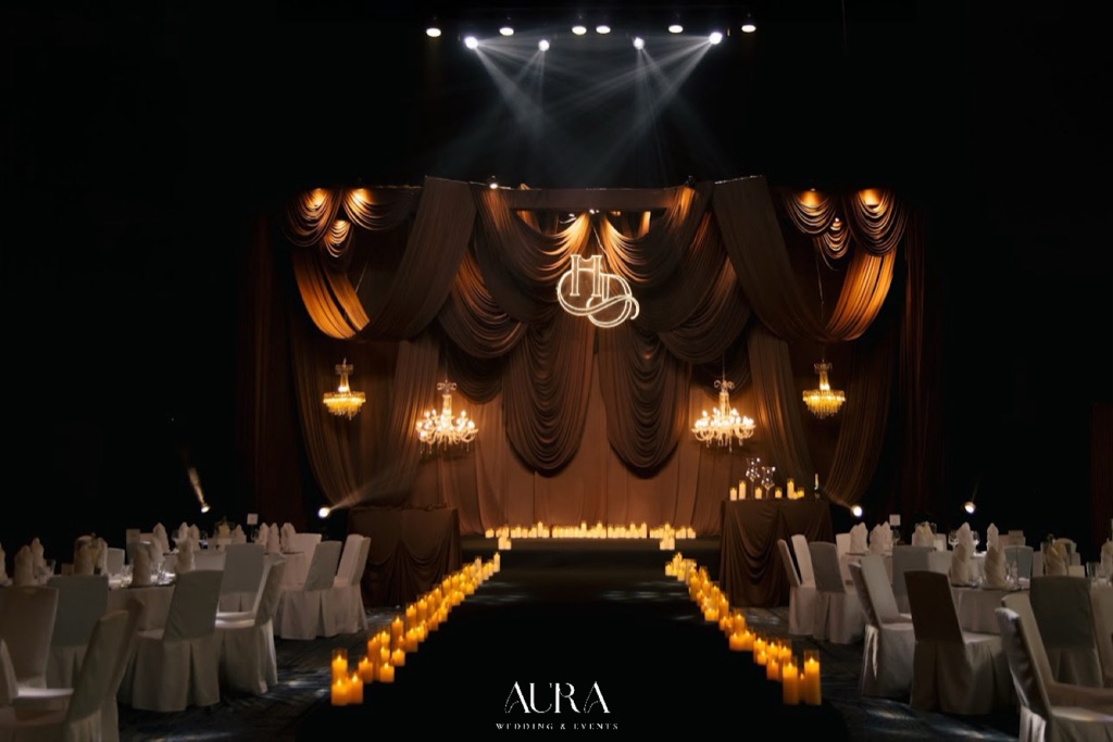Toàn cảnh lễ đường The Amber Hour: sân khấu rèm nâu socola xếp nếp, bốn đèn chùm pha lê, monogram HD và lối đi viền nến giữa các bàn tiệc