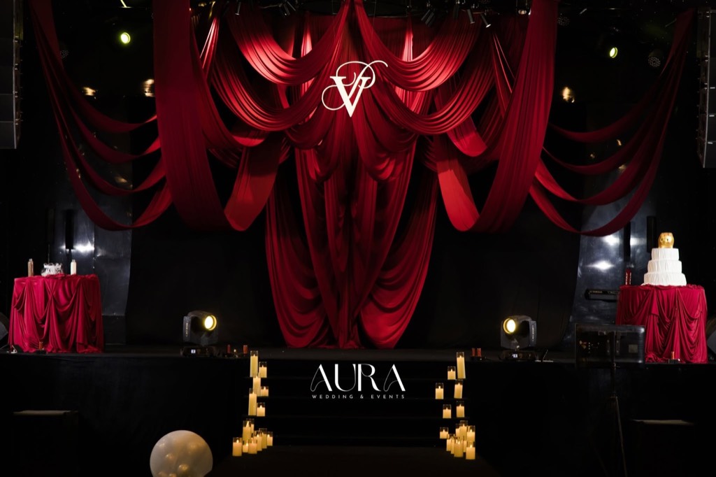 Sân khấu lụa đỏ thẫm buông rủ nhiều tầng với monogram VP, bậc thang viền nến, bàn bánh cưới và bàn phủ khăn đỏ hai bên