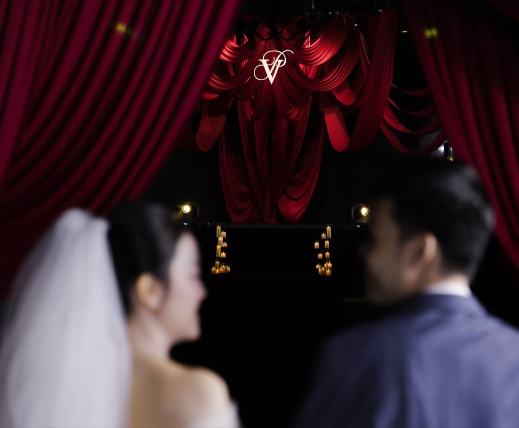 Cô dâu chú rể mờ ở tiền cảnh, hướng về sân khấu lụa đỏ và monogram VP