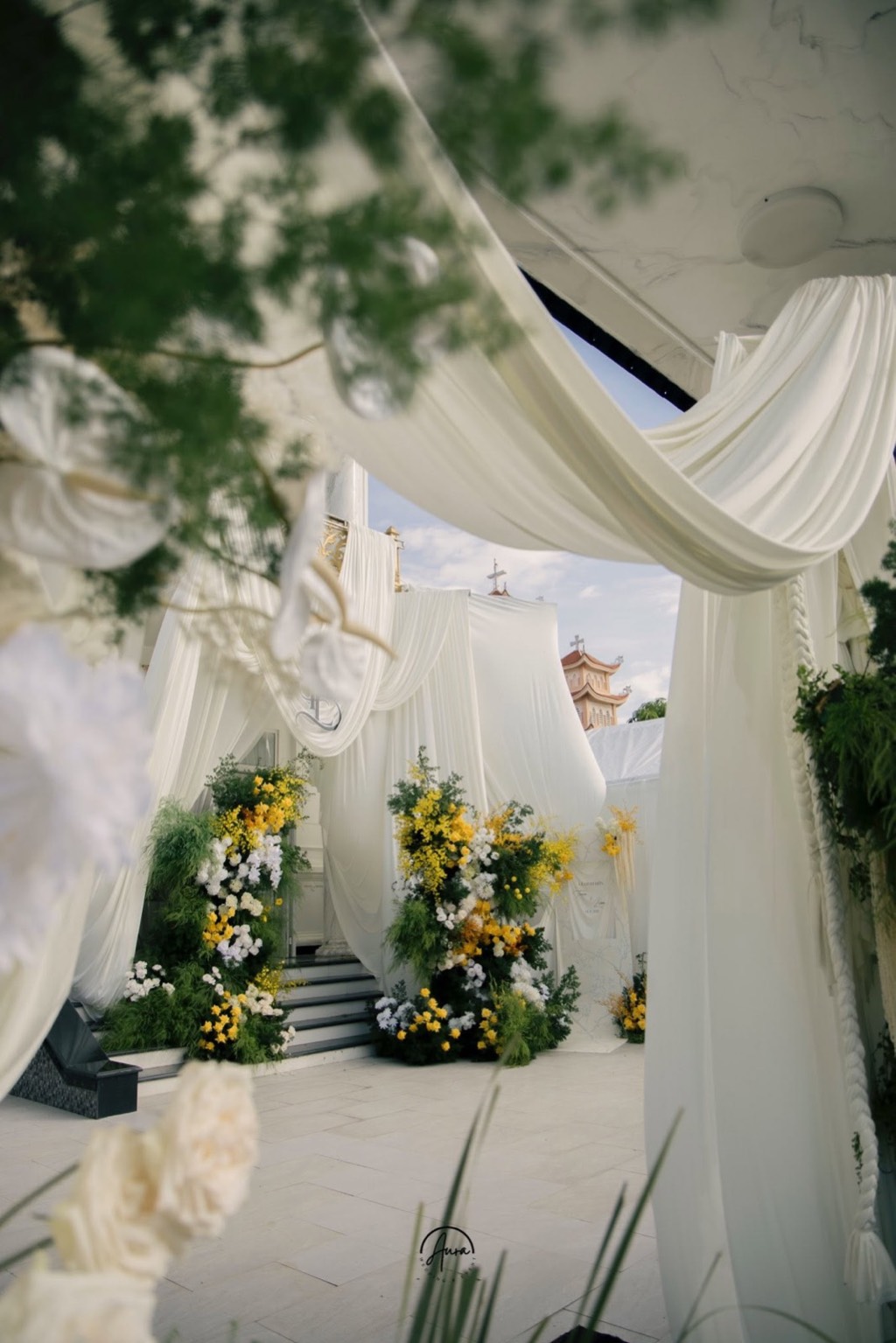 Cổng lễ đính hôn nhìn qua những lớp lụa trắng rủ, hai khối hoa vàng và trắng dưới bầu trời xanh