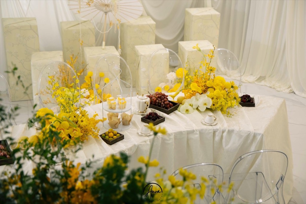 Bàn trà đính hôn với ấm sứ trắng, nho, bánh và hoa vàng, phía sau là các khối hộp bọc giấy hoa văn và dù giấy