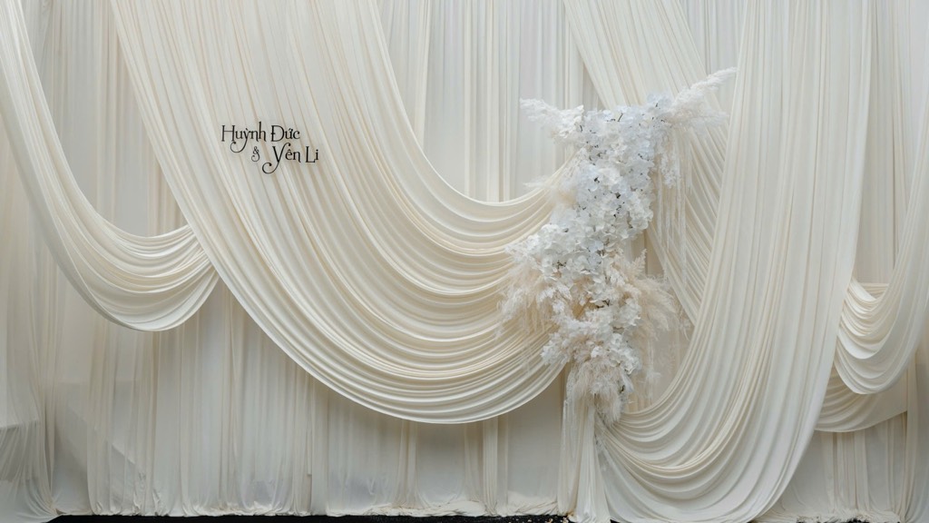 Backdrop chụp ảnh vải ngà buông võng nhiều tầng, bảng tên cặp đôi và cụm hoa trắng cùng cỏ lau